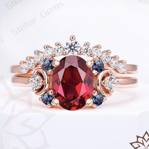 Könnte beinhalten: Ein roségoldenes Ring-Set mit einem großen, oval geschliffenen, tiefroten Edelstein. Der Ring ist mit kleineren weißen und blauen Edelsteinen in einem Halo-Design verziert. Ein passendes Band ergänzt das Set.