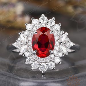 May include: A silver ring featuring a large, oval-cut, deep red gemstone at the center, surrounded by a halo of smaller, round, and marquise-cut white gemstones. The ring is set on a dark gray surface.