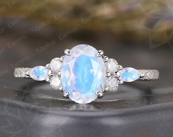 May include: A silver ring featuring a large, oval-cut, iridescent moonstone as the centerpiece. The moonstone is flanked by smaller, round, and pear-shaped gemstones. The band is adorned with additional small, clear stones. The ring is set against a dark background.
