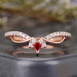 May include: Rose gold ring with a pear-shaped red gemstone center. The ring features a curved design with small, round white gemstones set along the band. The ring is set on a dark gray surface.