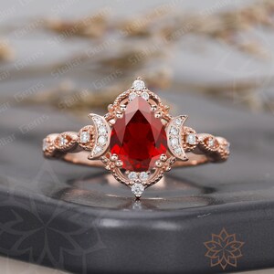 May include: A pear-shaped ruby engagement ring set in rose gold with a delicate filigree design and a halo of smaller diamonds. The ring features two crescent moon accents on either side of the centre stone.