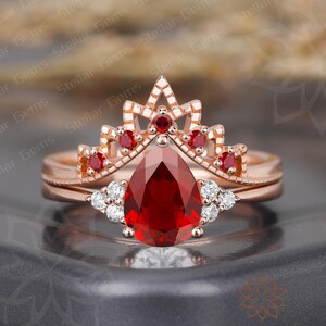 May include: A rose gold ring set featuring a pear-shaped, deep red gemstone. The ring is accented with small white gemstones and a crown-shaped top with red accents. The words "Stellar Gems" are visible in the image.