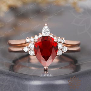 May include: A rose gold ring set featuring a pear-shaped, deep red gemstone as the focal point. The ring is accented with smaller, round, clear gemstones and a marquise-cut stone at the top. The set includes a matching band.