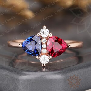 Könnte beinhalten: Ein Ring aus Roségold mit einem birnenförmigen blauen Saphir und einem birnenförmigen roten Rubin, die nebeneinander liegen. Eine vertikale Reihe aus vier kleinen runden weißen Diamanten trennt die Edelsteine, mit je einem größeren Diamanten an den Enden.