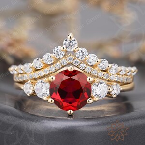 Puede incluir: Un juego de anillos dorados con una gema roja oscura, grande y redonda como punto focal. El anillo está adornado con gemas claras más pequeñas y un diseño en forma de corona. El conjunto incluye una banda a juego.