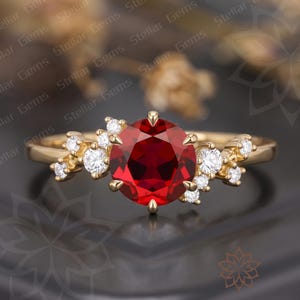 May include: A gold ring featuring a large, round, deep red center stone held by six prongs. The band is accented on both sides by a cluster of small, round, clear white stones arranged in a branching pattern along the gold metal shank.