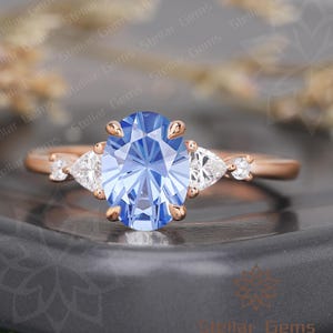 May include: Rose gold ring featuring a large, light blue oval-cut gemstone as the centerpiece. Flanking the center stone are two pear-shaped white gemstones, with smaller accent stones. The ring is a delicate piece of jewelry.