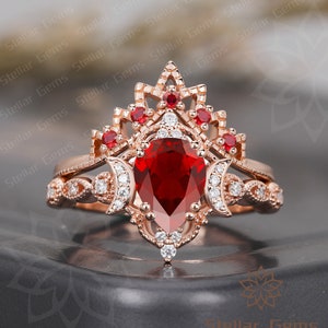 May include: A pear-shaped ruby engagement ring set in rose gold with a delicate crown-shaped halo and a matching band featuring a crescent moon design. The ring is adorned with smaller diamonds and rubies.