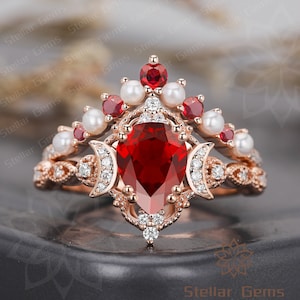 May include: A pear-shaped ruby gemstone ring set in rose gold with a delicate crown-like design featuring pearls and smaller red gemstones. The ring is accented with smaller diamonds.