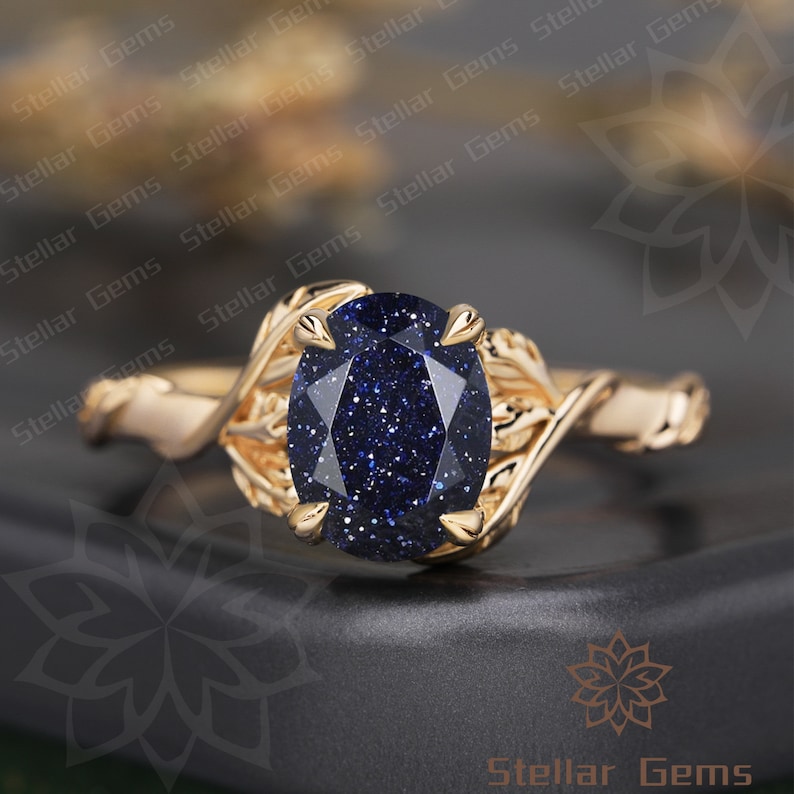 May include: A gold ring with a blue gemstone set in a leaf-shaped design. The gemstone is oval-shaped and has a sparkly, speckled texture.