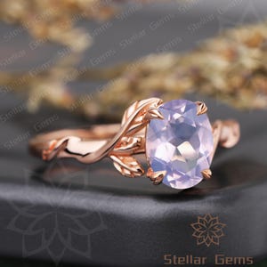 Puede incluir: Anillo de oro rosa con una piedra preciosa ovalada de color lavanda pálido. La banda tiene un diseño de hojas y vides. El anillo está sobre un fondo oscuro con las palabras "Stellar Gems".