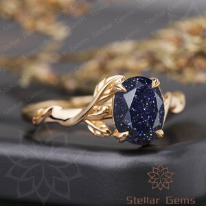 May include: A gold ring with a blue gemstone and leaf details. The ring is set on a black surface.
