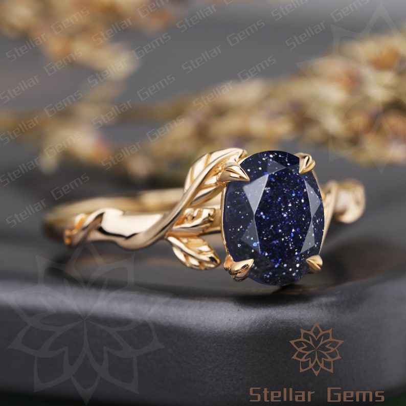 May include: A gold ring with a blue gemstone and leaf details. The ring is set on a black surface.