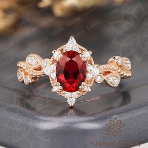 May include: A rose gold ring featuring a large oval-cut, deep red gemstone at the center, surrounded by a halo of smaller, clear gemstones. The band is adorned with additional small gemstones and decorative detailing. The ring is set on a dark gray surface.