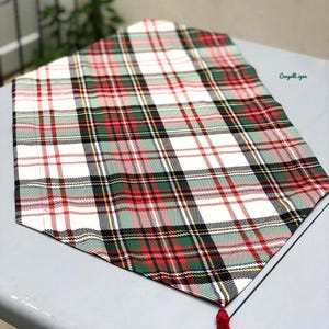 May include: A white table runner with a red, green, and black plaid pattern. The runner has a red tassel at the end.