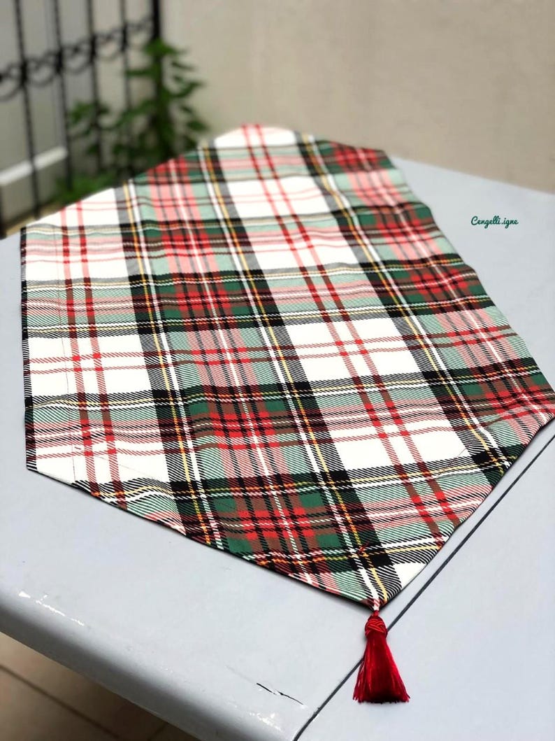May include: A white table runner with a red, green, and black plaid pattern. The runner has a red tassel at the end.
