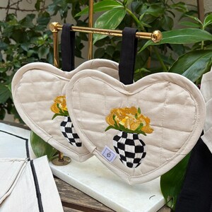 Puede incluir: Dos salvamanteles acolchados beige en forma de corazón con lazos de tela negra. Cada salvamanteles presenta un ramo de flores amarillas bordadas en un jarrón a cuadros blanco y negro. Los salvamanteles cuelgan de un estante dorado.