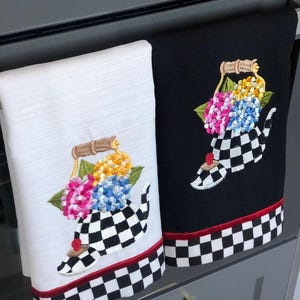 May include: Two kitchen towels, one white and one black, are displayed. Each towel features an embroidered teapot design with colorful flowers and a checkered pattern. A red border accents the bottom.