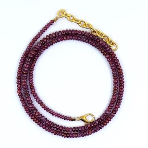 May include: A layered necklace composed of small, deep red beads. The necklace features a gold-colored chain and clasp. The beads are arranged in multiple strands, creating a full and elegant look. The necklace is displayed against a white background.