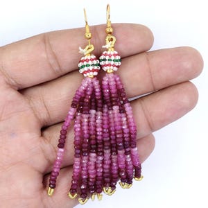 Könnte beinhalten: Ein Paar Hängeohrringe mit goldfarbenen Haken und kugelförmigen Perlen mit weißen, roten und grünen Mustern. Jeder Ohrring hat mehrere lange Stränge aus facettierten rosa und magentafarbenen Edelsteinperlen, die herabhängen.