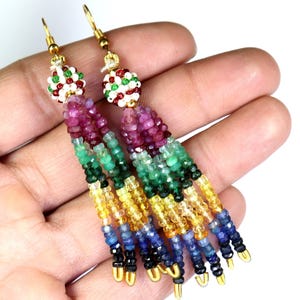 以下が含まれることがあります： カラフルな宝石の長いぶら下がりイヤリングのペア。イヤリングはルビー、エメラルド、サファイア、シトリンなど、さまざまな宝石で作られています。イヤリングは金色のワイヤーに通されており、上部に小さな丸い白いビーズが付いています。