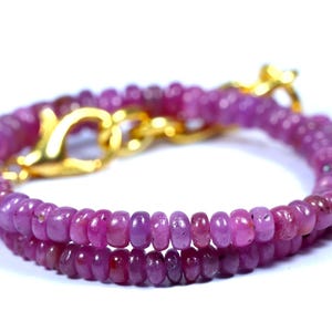 May include: A double-strand bracelet featuring small, round, pink gemstones. The bracelet is secured with a gold-colored clasp. The gemstones are a deep pink color, and the bracelet is displayed against a white background.
