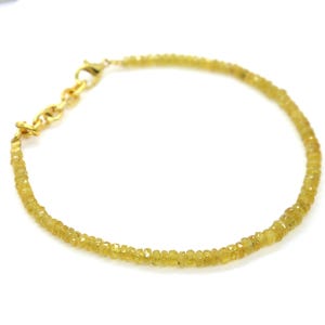Op de afbeelding: Een delicate armband met kleine, gefacetteerde gele kralen die aan elkaar zijn geregen. De armband heeft een goudkleurige sluiting en ketting. De kralen zijn lichtgeel van kleur en lijken edelstenen te zijn. De armband wordt op een witte achtergrond getoond.