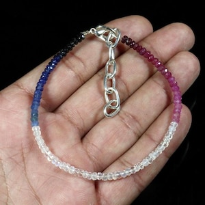 May include: A beaded bracelet featuring a gradient of faceted gemstones transitioning from deep black and blue to vibrant pink and clear white beads. The bracelet includes a silver-toned metal lobster clasp and an adjustable chain link extender.
