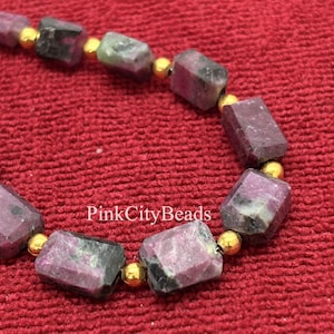 Puede incluir: Primer plano de un collar de cuentas con piedras preciosas rectangulares, facetadas, rosas y grises. Las cuentas están separadas por pequeñas cuentas doradas. El collar se muestra sobre un fondo rojo, con el texto "PinkCityBeads".