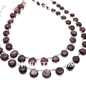 Pode incluir: Duas fileiras de contas de pedras preciosas roxas escuras. As contas são planas, arredondadas e enfiadas em um fio prateado. As contas têm um acabamento polido e brilhante e variam ligeiramente na cor, variando do bordô escuro a tons de roxo mais claros.