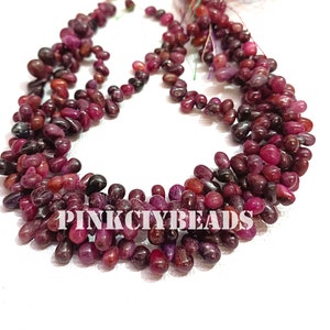 Op de afbeelding: Close-up van strengen dieprode en bordeauxrode druppelvormige kralen. De kralen zijn gegroepeerd, waardoor een textuur ontstaat. De tekst "PINK CITY BEADS" is zichtbaar onderaan de afbeelding.