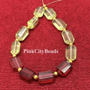 Pode incluir: Um fio de contas retangulares facetadas dispostas em forma de lágrima, com pedras de vidro translúcido em verde claro e vermelho escuro, separadas por pequenas contas espaçadoras de metal dourado. O texto PinkCityBeads está impresso em branco.