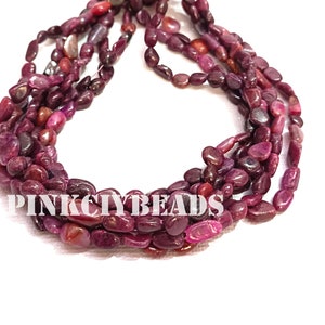 Puede incluir: Múltiples hebras de cuentas de rubí ovaladas pulidas en tonos rojos y rosas. Las cuentas están ensartadas, creando un efecto en capas. El texto "PINK CITY BEADS" es visible en la parte inferior de la imagen.