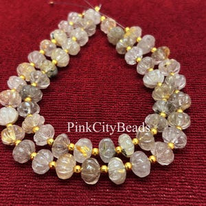 May include: Strand of clear, faceted beads with gold accents. The beads have a pumpkin shape and are strung together with gold-colored string. The beads are on a red background. The text "PinkCityBeads" is visible.
