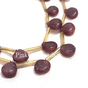 May include: Close-up of a strand of beads. The beads are a deep burgundy colour, with a teardrop shape. They are strung on gold-coloured metal rods. The beads are arranged in a repeating pattern. The background is white.