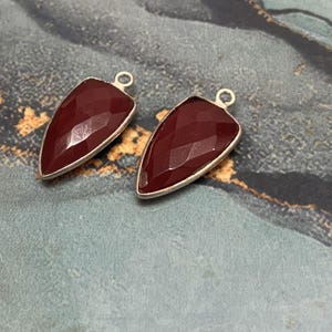 May include: Two maroon-colored, faceted gemstone pendants with a silver-colored metal border. Each pendant has a triangular shape with a small loop at the top for hanging. The gemstones have a polished, reflective surface.