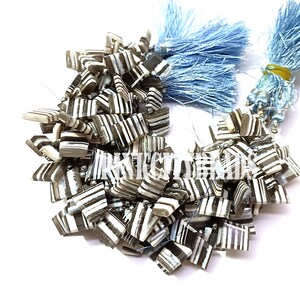 May include: A collection of square beads with alternating stripes of white and dark brown. The beads are strung together and accented with light blue tassels. The beads are a neutral color and could be used for jewelry making.