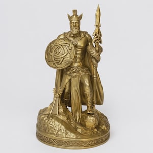 May include: A gold-toned statue of a bearded warrior wearing a winged helmet and a cape. The figure holds a tall spear in one hand and a round shield with an intricate triquetra knot design in the other, standing on a base with a small pyramid.