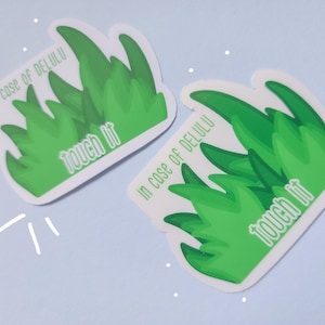 Op de afbeelding: Twee stickers met een groen grasontwerp en de tekst "In case of Delulu, touch it". De stickers hebben een witte rand en staan op een lichtblauwe achtergrond met witte steraccenten.