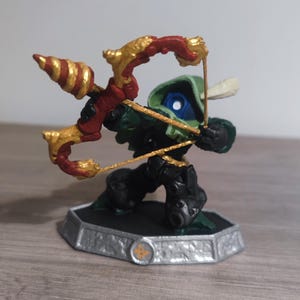 May include: A detailed figurine of a Skylanders character. The figure is mainly black and green, with a red and gold bow and arrow. The character is posed on a silver base with a gold emblem. The figurine is likely made of plastic.