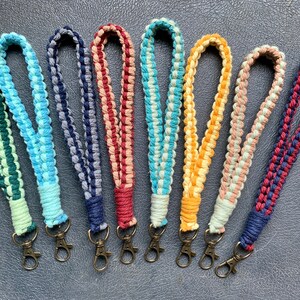 May include: Eight colorful braided keychains with metal key rings. The keychains are made of woven cotton cord in various colors, including green, blue, gray, red, pink, yellow, and beige.
