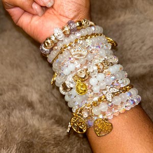 May include: A stack of bracelets with white beads, gold charms, and clear crystals. The charms include a crown, a heart, and a tree of life.