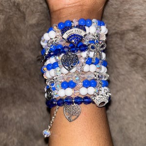Peut inclure: Plusieurs bracelets de perles bleues, blanches et transparentes. Les bracelets sont ornés de breloques, dont une couronne, un arbre de vie et un pendentif avec le texte "Finer 1920".