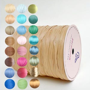 May include: A large spool of natural raffia ribbon with a variety of colored raffia samples. The ribbon is a light beige color, and the samples are arranged in a grid, showcasing various shades of blue, green, pink, and brown.