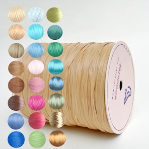 May include: A large spool of beige raffia ribbon with a flat, textured surface. The spool features a white circular end cap with the text ROOKIE and a small bird icon. To the left, a grid of twenty-four circular swatches displays various colors.
