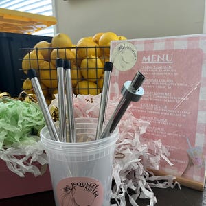 K&ouml;nnte beinhalten: Ein durchsichtiger Plastikbecher gef&uuml;llt mit wiederverwendbaren Edelstahlstrohhalmen. Der Becher hat einen rosa-wei&szlig;en Aufkleber mit den Worten "Squeeze Sister" und einem Zitronendesign. Der Becher steht auf einem Tisch mit einer rosa-wei&szlig;en karierten Speisekarte dahinter. Die Speisekarte listet verschiedene Limonade-Geschmacksrichtungen und Preise auf.