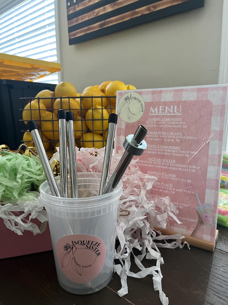 K&ouml;nnte beinhalten: Eine Nahaufnahme eines durchsichtigen Plastikbeh&auml;lters, der mit Edelstahl-Trinkhalmen gef&uuml;llt ist. Der Beh&auml;lter hat einen rosa-wei&szlig;en Aufkleber mit den Worten "Squeeze Sister Lemonade". Der Beh&auml;lter steht auf einem Tisch mit einer rosa-wei&szlig;en karierten Speisekarte dahinter. Die Speisekarte listet verschiedene Limonade-Geschmacksrichtungen auf, darunter "Classic Lemonade", "Mon Pina Colada", "Sister Strawberry" und "Ocean Water".