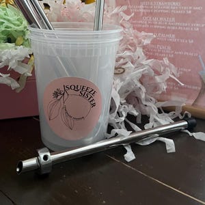 K&ouml;nnte beinhalten: Ein durchsichtiger Plastikbecher mit einem rosa und wei&szlig;en Aufkleber, auf dem "Squeeze Sister Lemonade Vendor" steht, und einem silbernen Metallstrohhalm.