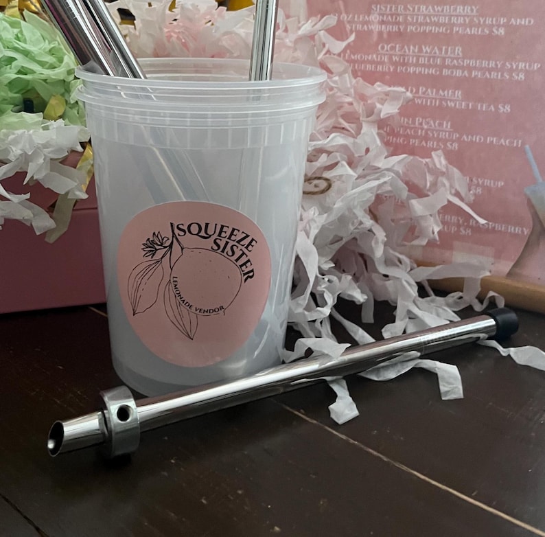 K&ouml;nnte beinhalten: Ein durchsichtiger Plastikbecher mit einem rosa und wei&szlig;en Aufkleber, auf dem "Squeeze Sister Lemonade Vendor" steht, und einem silbernen Metallstrohhalm.