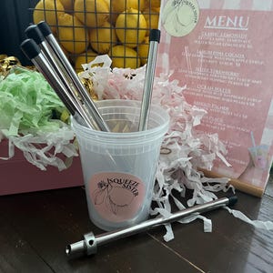 K&ouml;nnte beinhalten: Nahaufnahme eines durchsichtigen Plastikbechers mit einem rosa-wei&szlig;en Logo, auf dem "Squeeze Sister Lemonade & Tea" steht. Der Becher ist mit bunten Papierschnitzeln und vier wiederverwendbaren Edelstahlstrohhalmen gef&uuml;llt. Ein Edelstahlstrohhalm mit schwarzer Spitze liegt auf einem Holztisch.